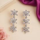 A pair of silver-toned dangle earrings with three stacked starburst motifs, each adorned with clear crystals, displayed on a light beige surface with decorative pink and purple floral/satin props.