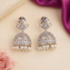 A pair of silver-toned Indian jhumka earrings adorned with clear crystals and dangling white pearls, displayed on a beige surface with decorative pink rose and purple fabric props.