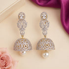 A pair of ornate silver-toned jhumka earrings embellished with clear crystals and gold accents, each hanging from a round crystal stud and featuring a dangling pearl-like bell, displayed on a beige surface with a pink rose and purple fabric in the background.