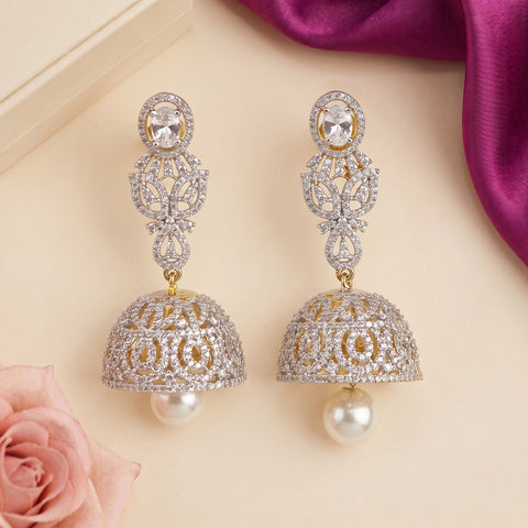 A pair of ornate silver-toned jhumka earrings embellished with clear crystals and gold accents, each hanging from a round crystal stud and featuring a dangling pearl-like bell, displayed on a beige surface with a pink rose and purple fabric in the background.