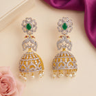A pair of ornate gold and silver-toned jhumka earrings decorated with green stones, pavé-set clear stones, and dangling pearl-like beads, arranged on a beige surface with a pink rose and purple fabric accents.