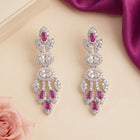 A pair of silver-toned drop earrings adorned with clear crystals and pink gemstones, displayed on a beige surface with a pink rose and purple fabric in the background.