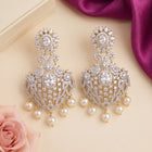 A pair of ornate gold-toned chandelier earrings adorned with clear crystals and white faux pearls, displayed on a beige surface with a pink rose and purple satin fabric in the background.