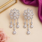 A pair of silver-toned statement drop earrings adorned with clear crystals, arranged on a beige surface with decorative pink fabric and a purple satin corner.