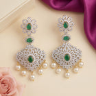 A pair of ornate Indian-style chandelier earrings with green emerald-cut stones, silver pavé settings, and dangling white pearls, styled on a beige surface with a pink rose and purple satin ribbon.