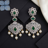 A pair of ornate silver-toned dangle earrings with green stone centers, pavé-set clear stones, and dangling pearl-like beads, arranged on a dark background with decorative fabric and a blue flower prop.