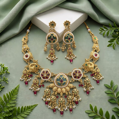 An ornate gold-toned traditional jewelry set displayed on a sage green fabric background, including a necklace, matching earrings, and a central decorative block. The jewelry features intricate filigree, green enamel details, red stone accents, and white pearl-like beads, with green leaves placed around the arrangement.