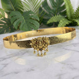 An ornate gold-toned metal choker belt featuring intricate floral engravings and a central Ganesha pendant with dangling bells, displayed on a white marble surface with green tropical leaves in the background.