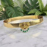 A gold-toned traditional Indian choker necklace with intricate filigree work, featuring a central Ganesha motif embellished with small red stones and hanging green bead drops, displayed on a white marble surface with green foliage in the background.