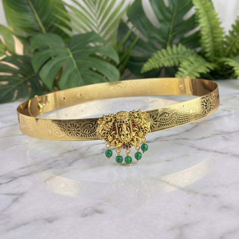 A gold-toned traditional Indian choker necklace with intricate filigree work, featuring a central Ganesha motif embellished with small red stones and hanging green bead drops, displayed on a white marble surface with green foliage in the background.