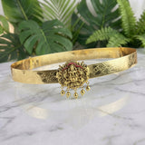 An ornate gold-toned traditional bangle with intricate engravings and a central deity pendant featuring red enamel details, displayed on a white marble surface with green foliage in the background.