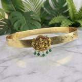 An ornate gold-toned traditional headband or choker with engraved patterns, a central deity motif, and dangling green bead accents, displayed on a white marble surface with green foliage in the background.