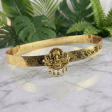 An ornate gold-toned traditional Indian choker necklace with a central deity pendant and dangling bells, displayed on a white marble surface with green plants in the background.
