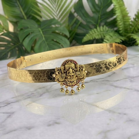 An ornate gold-toned traditional Indian choker necklace with a central deity pendant and dangling bells, displayed on a white marble surface with green plants in the background.