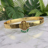 An ornate gold-toned traditional Indian choker belt with intricate filigree work, a central deity pendant, and dangling green bead drops, displayed on a white marble surface with green foliage in the background.