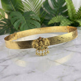 An ornate gold-toned traditional Indian choker bangle with intricate engravings and a central deity pendant with hanging bells, displayed on a white marble surface with green foliage in the background.