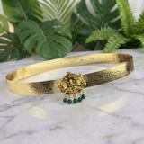 An ornate gold-toned traditional choker belt with intricate engraved patterns and a central deity pendant featuring green bead drops, displayed on a white marble surface with green foliage in the background.