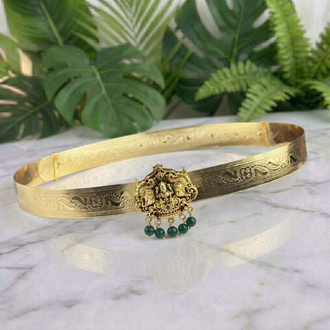 An ornate gold-toned traditional choker belt with intricate engraved patterns and a central deity pendant featuring green bead drops, displayed on a white marble surface with green foliage in the background.