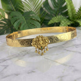 An ornate gold-toned traditional Indian bangle bracelet with intricate floral engravings and a central Ganesha pendant with dangling bells, displayed on a white marble surface with green foliage in the background.