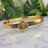 An ornate gold-toned traditional Indian bangle bracelet with intricate floral engravings and a central hanging Ganesha pendant, displayed on a white marble surface with green tropical leaves in the background.