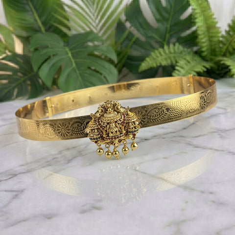 An ornate gold-toned traditional Indian bangle bracelet with intricate floral engravings and a central hanging Ganesha pendant, displayed on a white marble surface with green tropical leaves in the background.