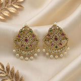 A pair of ornate gold-toned Indian jhumka earrings embellished with green and pink stones and dangling white faux pearls, displayed on a cream satin fabric background with decorative gold leaf elements.