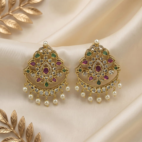 A pair of ornate gold-toned Indian jhumka earrings embellished with green and pink stones and dangling white faux pearls, displayed on a cream satin fabric background with decorative gold leaf elements.