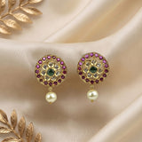 A pair of gold-plated Indian Kundan earrings with a central green stone, surrounded by pink and clear stones, with a dangling white pearl, displayed on a beige satin fabric background with gold decorative leaves.