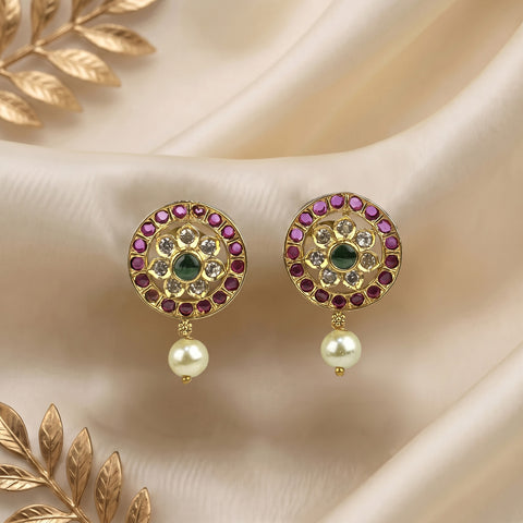 A pair of gold-plated Indian Kundan earrings with a central green stone, surrounded by pink and clear stones, with a dangling white pearl, displayed on a beige satin fabric background with gold decorative leaves.