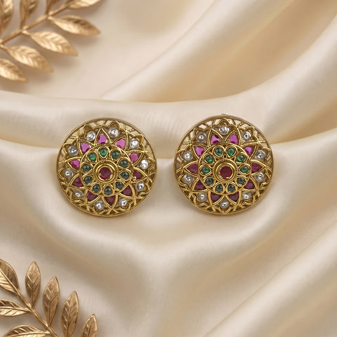 A pair of gold-plated Indian Kundan earrings with intricate mandala designs, featuring pink, green, and white stones, displayed on a cream satin fabric with gold leaf accents.