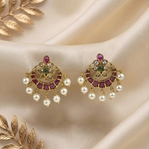 A pair of gold-toned Indian jhumka earrings decorated with pinkish-red stones, green stones, and dangling white faux pearls, displayed on a draped cream satin fabric with gold leaf accents.