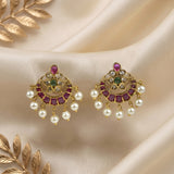 A pair of gold-toned chandelier earrings with pinkish-red stones, green stones, and dangling white pearls, displayed on draped cream satin fabric with decorative gold leaf elements.