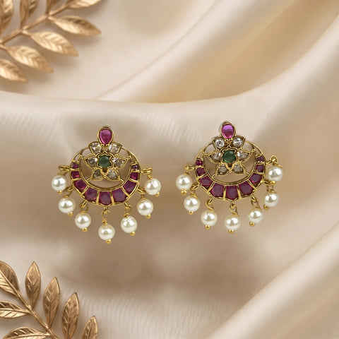 A pair of gold-toned chandelier earrings with pinkish-red stones, green stones, and dangling white pearls, displayed on draped cream satin fabric with decorative gold leaf elements.