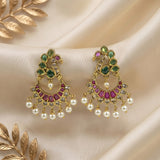A pair of gold-plated Indian chandbali earrings decorated with green and pink stones and dangling white faux pearls, displayed on a draped cream satin fabric with gold leaf accents.