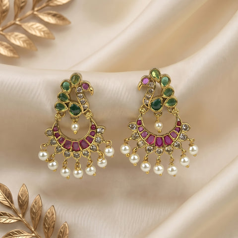 A pair of ornate gold-toned chandelier earrings decorated with green stones, pink stones, and dangling white pearls, displayed on a cream satin fabric with gold leaf accents.
