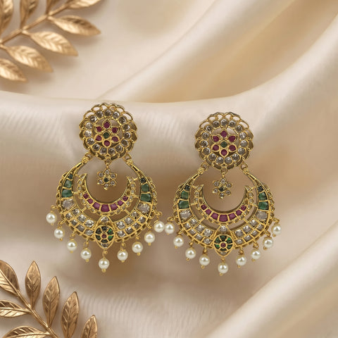 A pair of ornate gold-toned Indian chandelier earrings decorated with green and pink enamel, clear stones, and dangling white pearls, displayed on draped cream satin fabric with gold leaf accents.