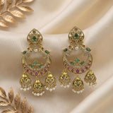 A pair of ornate gold-toned Indian jhumka earrings decorated with green stones, pink stones, and small white pearl beads, displayed on draped cream satin fabric with decorative gold leaf elements.