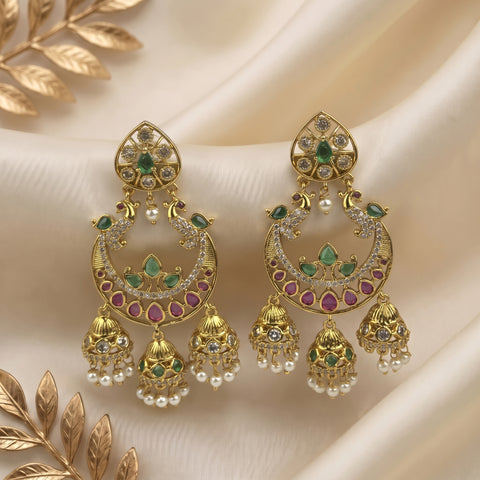 A pair of ornate gold-toned Indian jhumka earrings decorated with green stones, pink stones, and small white pearl beads, displayed on draped cream satin fabric with decorative gold leaf elements.
