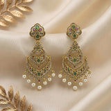 A pair of ornate gold-toned Indian chandelier earrings decorated with green and pink stones, clear crystals, and dangling white faux pearls, displayed on draped cream satin fabric with gold leaf accents.