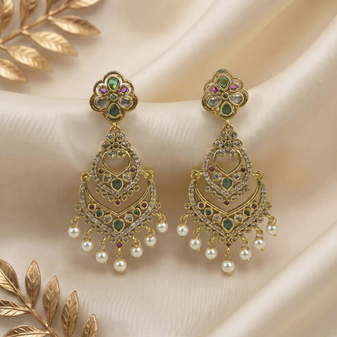 A pair of ornate gold-toned Indian chandelier earrings decorated with green and pink stones, clear crystals, and dangling white faux pearls, displayed on draped cream satin fabric with gold leaf accents.