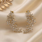 Gold crescent-shaped dangle earrings adorned with marquise and round clear crystals, arranged on draped cream satin fabric with a gold leaf prop in the corner.