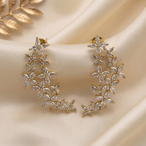 Gold crescent-shaped dangle earrings adorned with marquise and round clear crystals, arranged on draped cream satin fabric with a gold leaf prop in the corner.