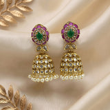 A pair of gold-plated Indian jhumka earrings with green stone centers, purple stone accents, and white pearl bead borders, displayed on a beige satin fabric background with decorative gold leaves.