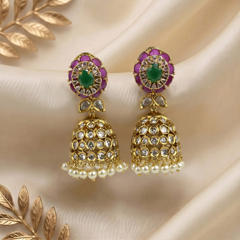 A pair of gold-plated Indian jhumka earrings with green stone centers, purple stone accents, and white pearl bead borders, displayed on a beige satin fabric background with decorative gold leaves.