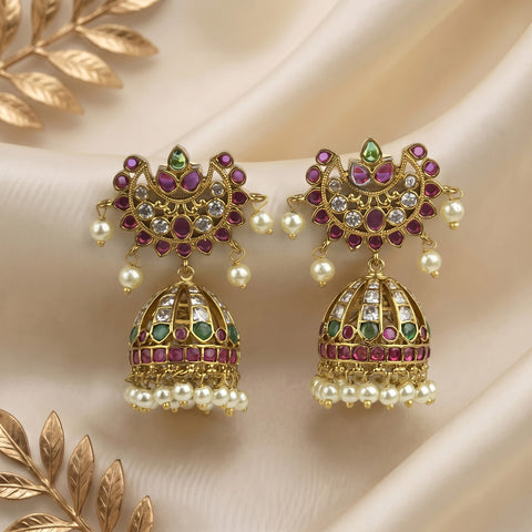 A pair of ornate gold-toned Indian jhumka earrings decorated with purple, green, and clear stone accents, hanging pearls, and a gold-toned beaded fringe, displayed on draped cream satin fabric with decorative gold leaves.