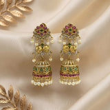 A pair of ornate gold-toned jhumka earrings decorated with pink gemstones, green stones, clear crystals, and white pearl beads, displayed on draped cream satin fabric with decorative gold leaf elements.