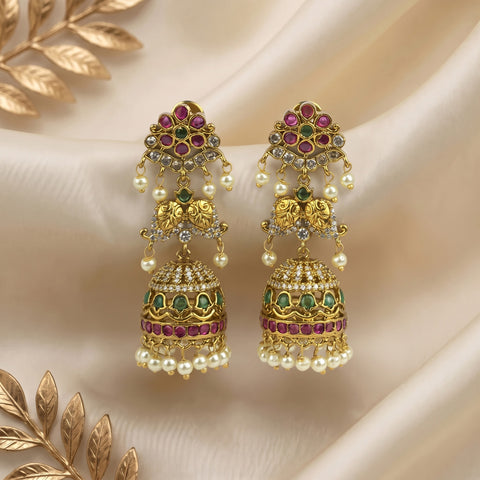 A pair of ornate gold-toned jhumka earrings decorated with pink gemstones, green stones, clear crystals, and white pearl beads, displayed on draped cream satin fabric with decorative gold leaf elements.