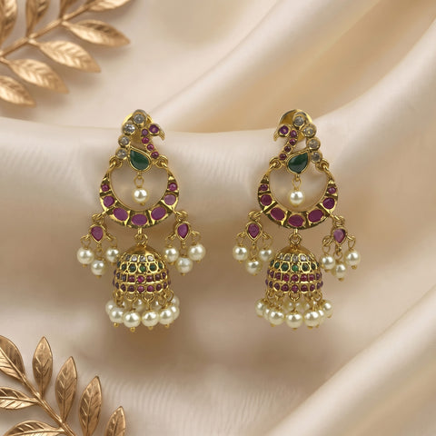 A pair of ornate gold-toned Jhumka earrings embellished with green and purple stones and white faux pearls, displayed on draped cream satin fabric with decorative gold leaf elements.