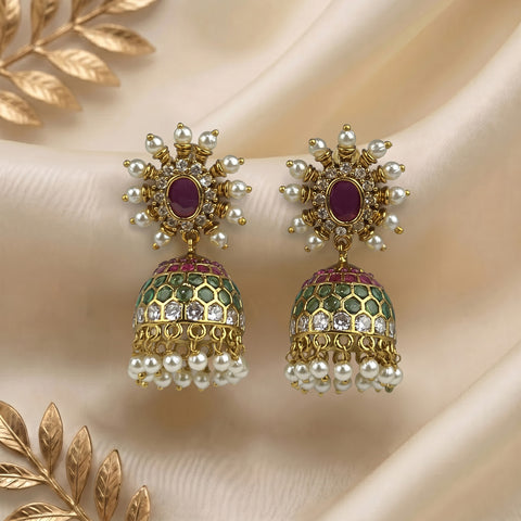 A pair of ornate Indian jhumka earrings with gold-plated frames, purple oval stones, green and white marbled enamel, and dangling white pearl beads, displayed on a beige satin fabric background with gold decorative leaves.