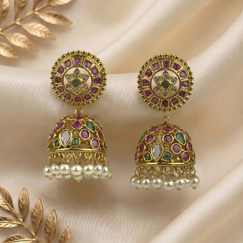 A pair of ornate gold-toned jhumka earrings with multicolored stone inlays, white pearl bead drops, and circular floral motifs, displayed on draped cream fabric with decorative gold leaves.